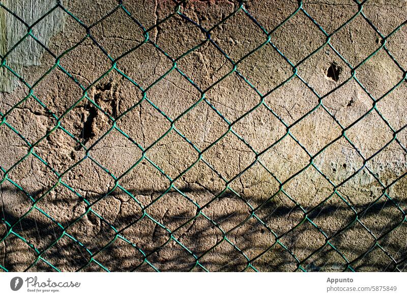 Wall, fence and time - details of an old wall with cracks and scar-like indentations behind a shadow-casting green wire mesh fence Fence Wall (barrier)