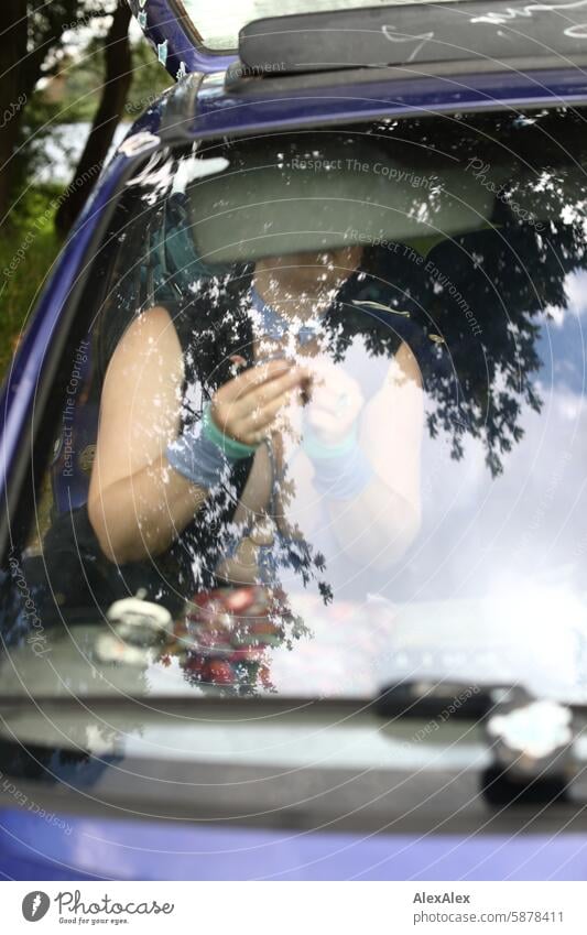 A woman with a large neckline is sitting in the front right-hand side of her car, putting on make-up and jewelry. You can't see her through the reflection in the windshield