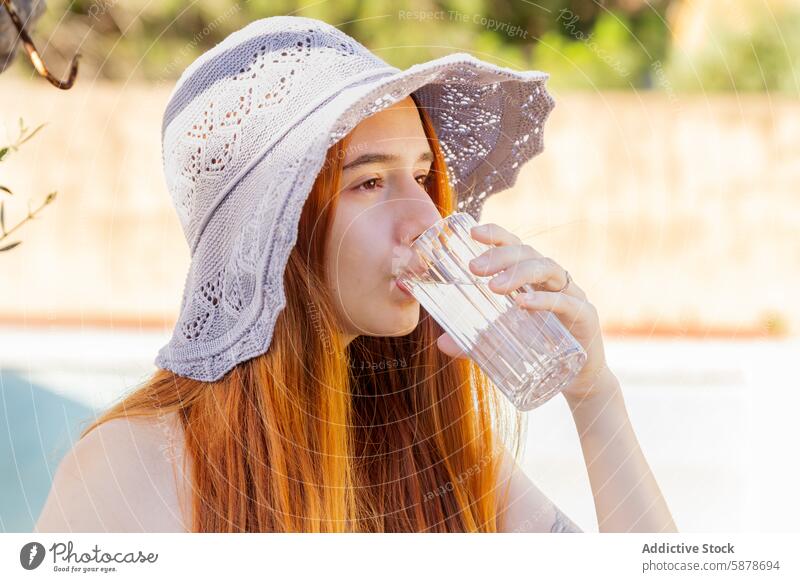 Young woman enjoys refreshment by the pool in summer water drink glass hat lace poolside red hair young outdoor sunny hydrate lifestyle leisure relaxation