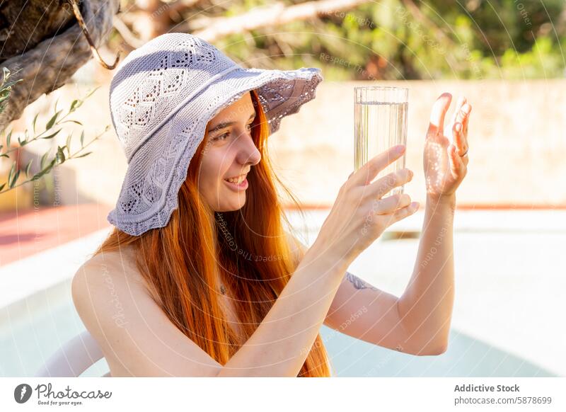 Young woman enjoying a glass of water by the pool summer hat sunshine relaxation outdoor happiness leisure lifestyle sunny redhead crochet drinking hydration