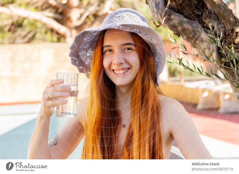 Young woman enjoying a sunny summer day by the pool hat water glass smile cheerful outdoor lifestyle refreshing relaxed poolside young redhead beverage drinking