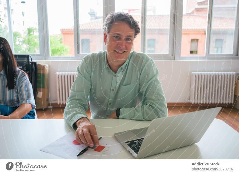 Mature businessman working at a laptop in a bright office mature professional paperwork sitting table focused approachable environment casual wear shirt green