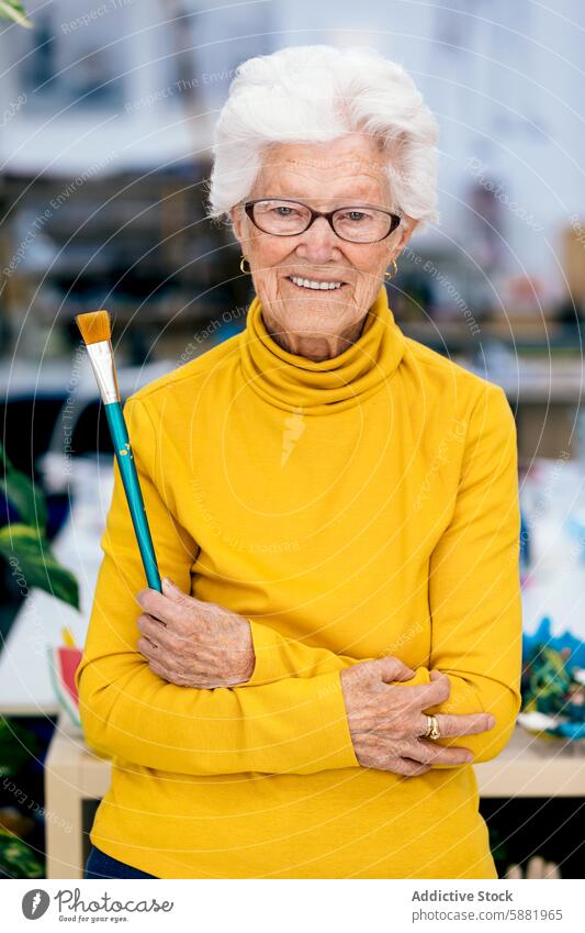 Elderly woman painter holding a brush in art studio elderly painting creative portrait cheerful mature senior artist hobby yellow turtleneck glasses workshop