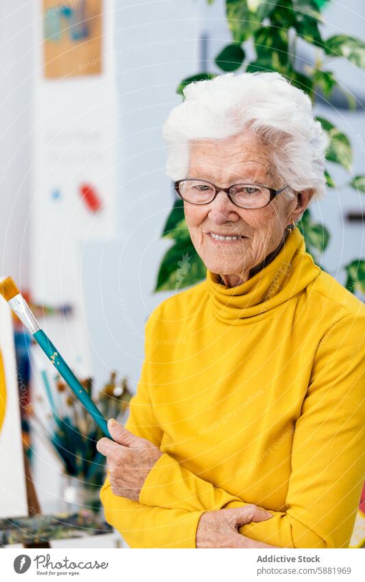 Elderly woman artist holding a paintbrush in a studio elderly senior mature painting cheerful yellow turtleneck supplies creativity hobby leisure crafting