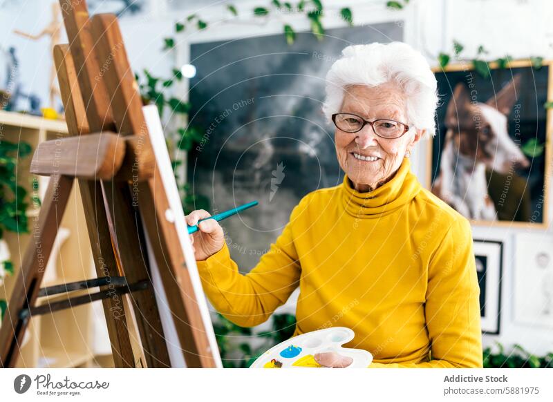 Elderly woman paints on canvas in a bright studio elderly senior mature painting art cheerful creative hobby painter brush palette artistic yellow turtleneck