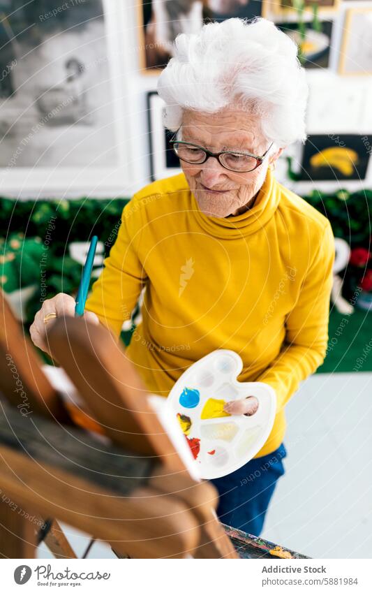 Elderly woman painting in her art studio elderly senior mature artist palette brush canvas creative hobby lifestyle glasses yellow top joyful expressive