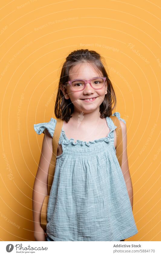 Smiling schoolgirl with glasses against a yellow background smile blue dress portrait studio child female education cheerful vibrant preschooler kindergarten