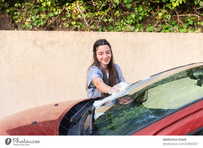 Young woman washing car windshield outdoors car wash cleaning smiling soap water squeegee vehicle car care manual labor hobby sunny day nature foliage hygiene