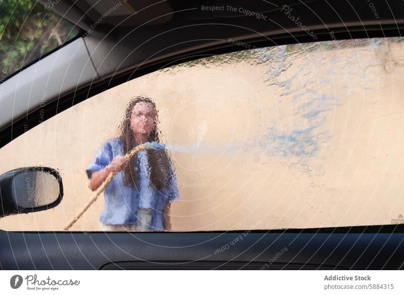 Unrecognizable woman washing car as seen through a wet window car wash cleaning vehicle hose water spray focus task not looking at camera car window steamy