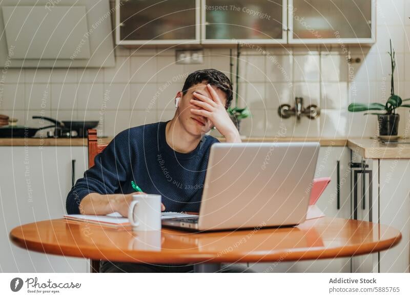 Frustrated young man working from home on a laptop stress overwhelmed remote work kitchen table coffee mug paper home office frustration young adult male