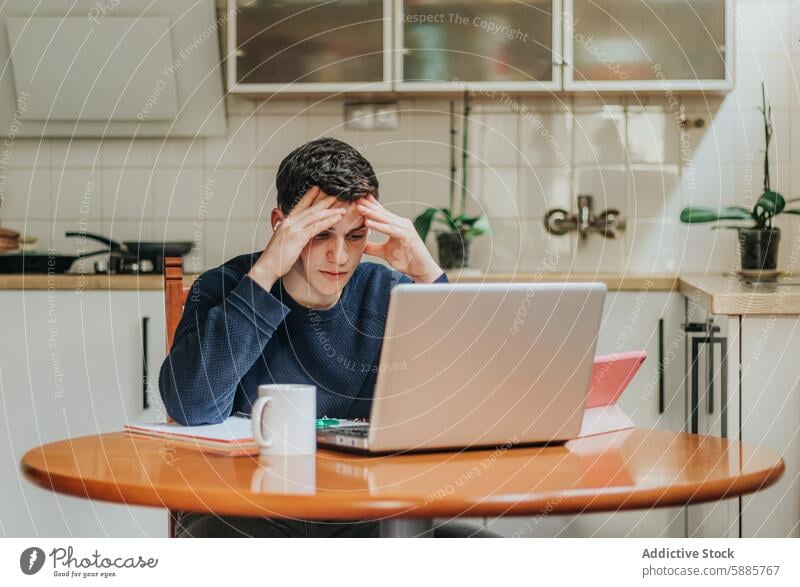 Young man stressed during a home study session laptop kitchen table work tired pressured focus anxiety education remote distance learning book notebook mug