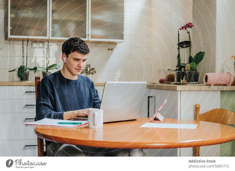 Young man working from home in a cozy kitchen setting laptop table coffee mug plant sunny focus technology wireless earbuds notebook pen wooden chair window