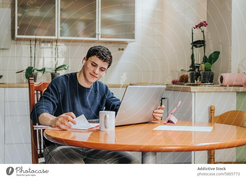 Young man studying with digital devices at home kitchen young laptop notebook smartphone table chair male adult student learning education technology indoor