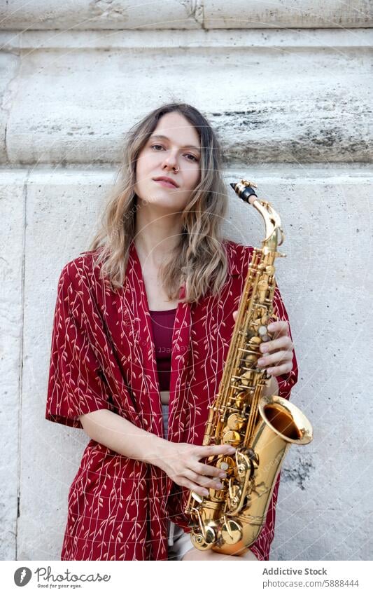 Woman holding saxophone against an old stone wall woman music musician instrument jazz female blazer patterned red aged calm gaze off-camera young stylish