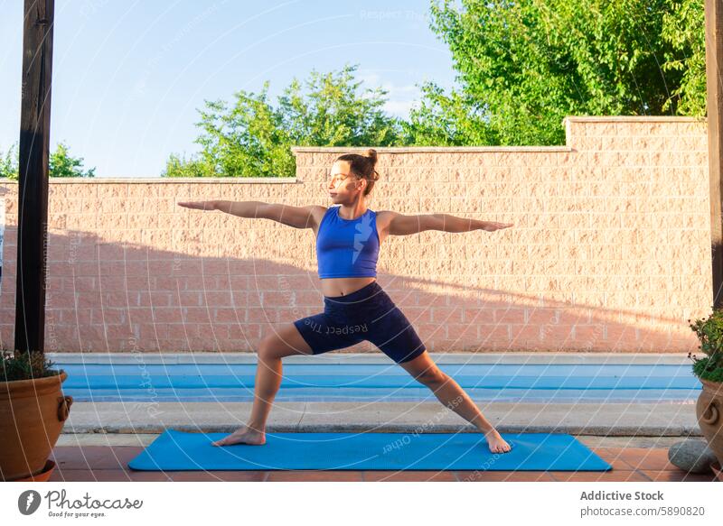 Woman practicing yoga beside a serene pool setting woman pose warrior ii mat tranquil sun evening greenery wall outdoors fitness health wellness exercise