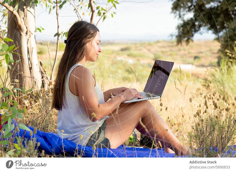 Young woman working on laptop in a rural setting outdoors nature grass tree field technology remote environment countryside summer relaxed focused sitting