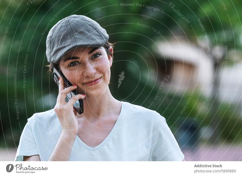 Woman in a cap speaking on a mobile phone outdoors woman smartphone phone call talk technology device gadget flat cap white shirt smiling looking at camera