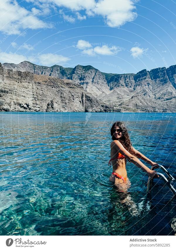 Young woman enjoys sunny beach day by mountainous coast summer holiday young bikini orange swim blue water clear sky smile vibrant nature outdoor sea vacation