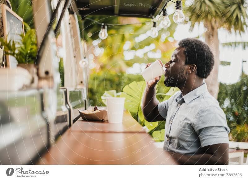 Young african man eating and drinking in food truck counter outdoor in city park - Focus on face bar beverage brazil buying cafeteria coffee customer delivery