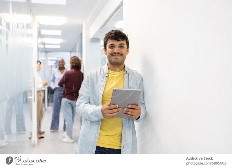 Smiling academic holding tablet in a busy corridor man casual cheerful school conversation colleague digital device attire education background professional