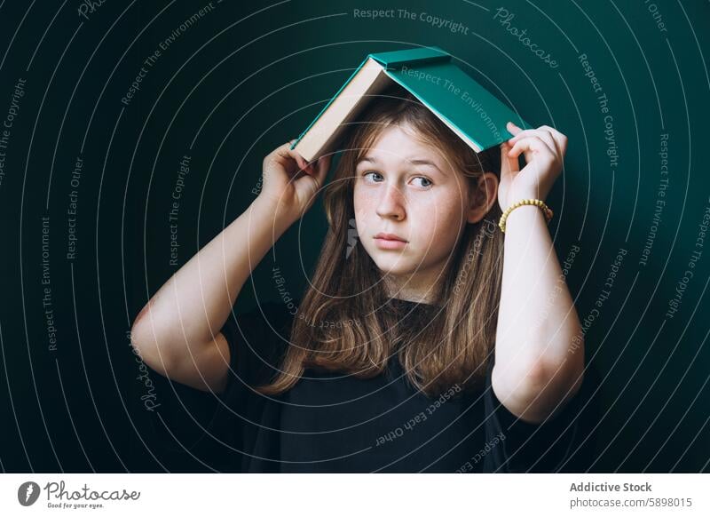 Young girl with book on head against dark backdrop student education back to school thoughtful portrait dark background contemplation schoolgirl young female