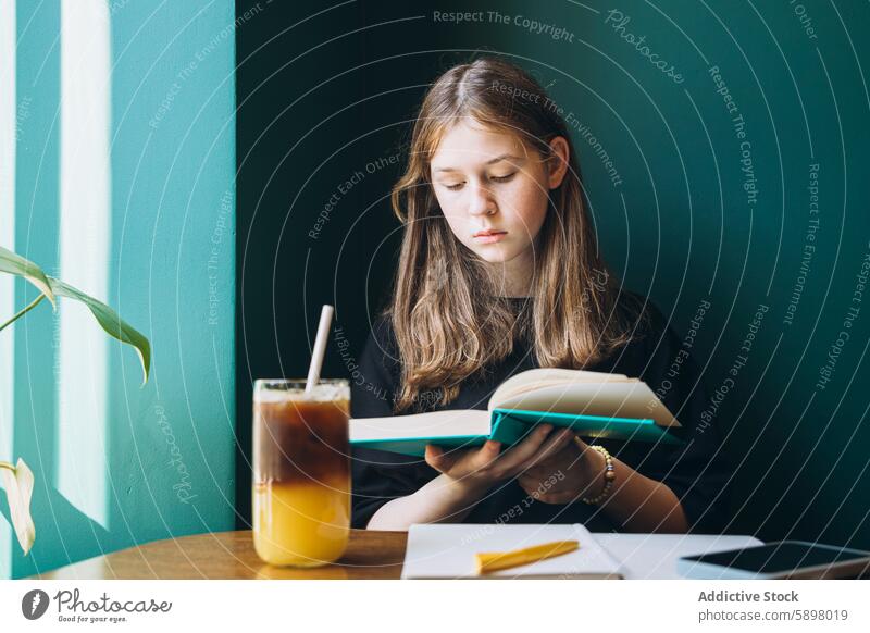 Young girl studying alone in a cafe with a smartphone nearby student female school book reading learning looking down table chair beverage drink cold drink cup