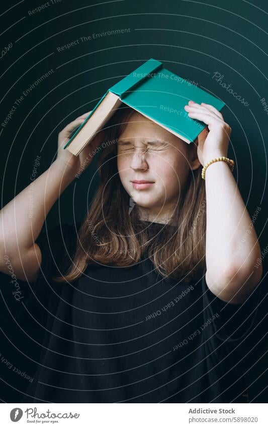 Young girl using book as a hat in a moment of frustration student stress education school overwhelmed dark background young female eyes closed expression strain