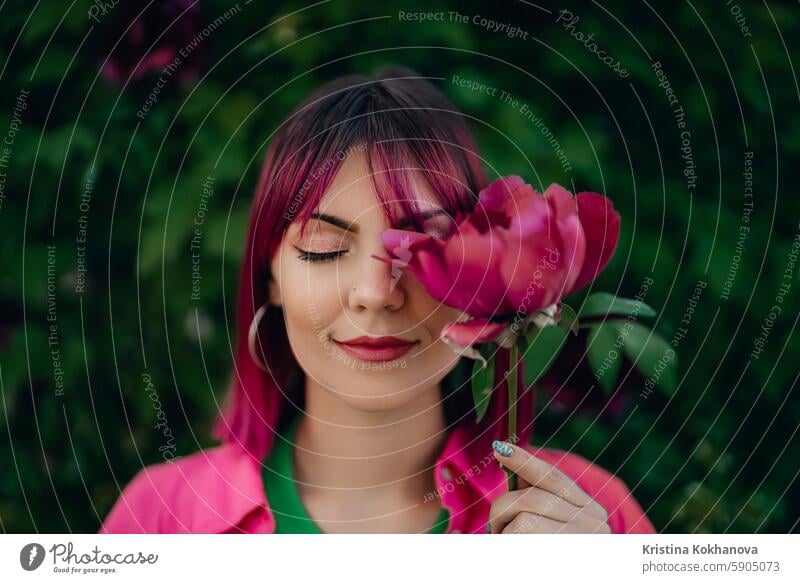 Portrait of real serene woman with vivid dyed pink fuchsia hairstyle. Peony flower, green nature backdrop. Trendy colorful hair, stylish haircut beautiful