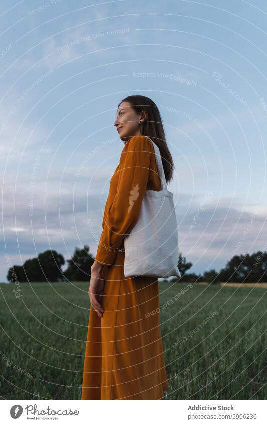 Woman in orange dress holding tote bag in a field woman nature outdoors sky gaze looking away green blue serene contemplative brunette summer cloth casual