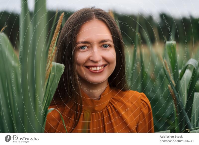 Young brunette woman looking at camera in a green field orange blouse smiling foliage nature outdoor female young cheerful eye contact portrait summer rural