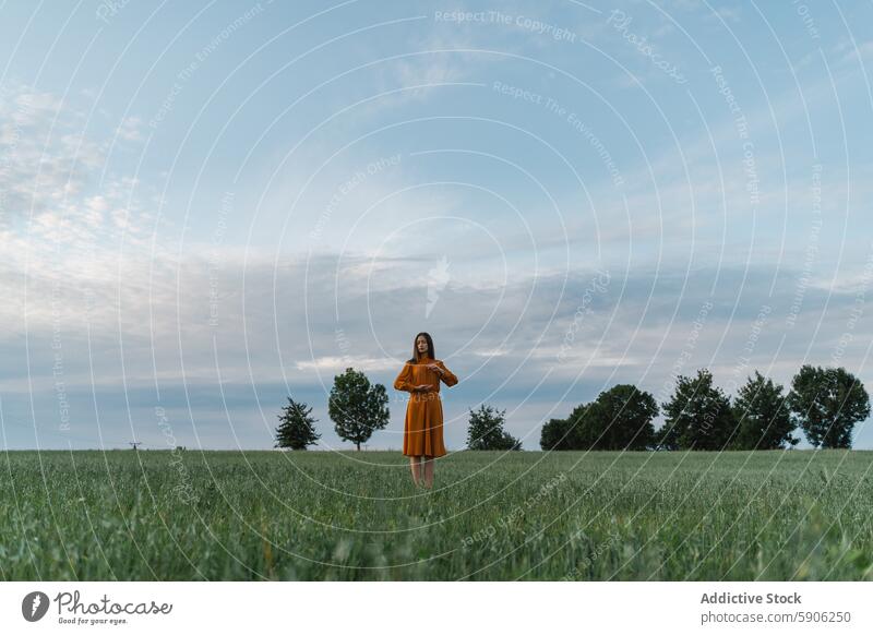 Woman in golden dress stands in lush green field under cloudy sky woman brunette tranquility nature landscape serene outdoor countryside solitude summer scenic