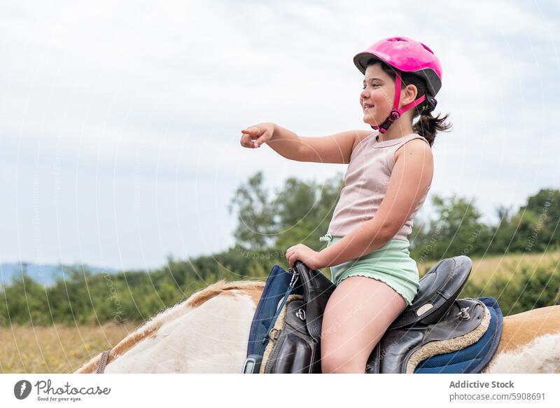 Young girl riding horse and pointing to the distance helmet horizon smile saddle equestrian adventure outdoor rural countryside nature child leisure activity