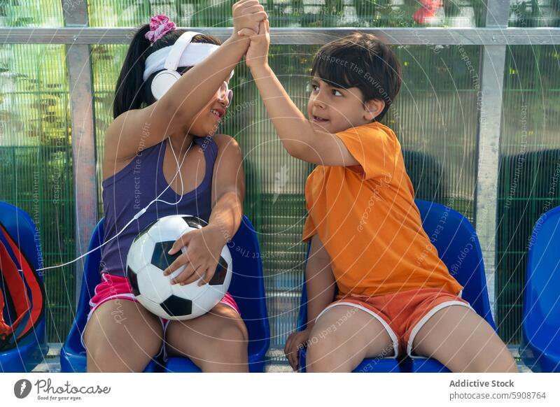 Two children enjoy a playful moment with a soccer ball sport interaction celebration high five bench outdoor fence sit summer smile casual friendship youthful