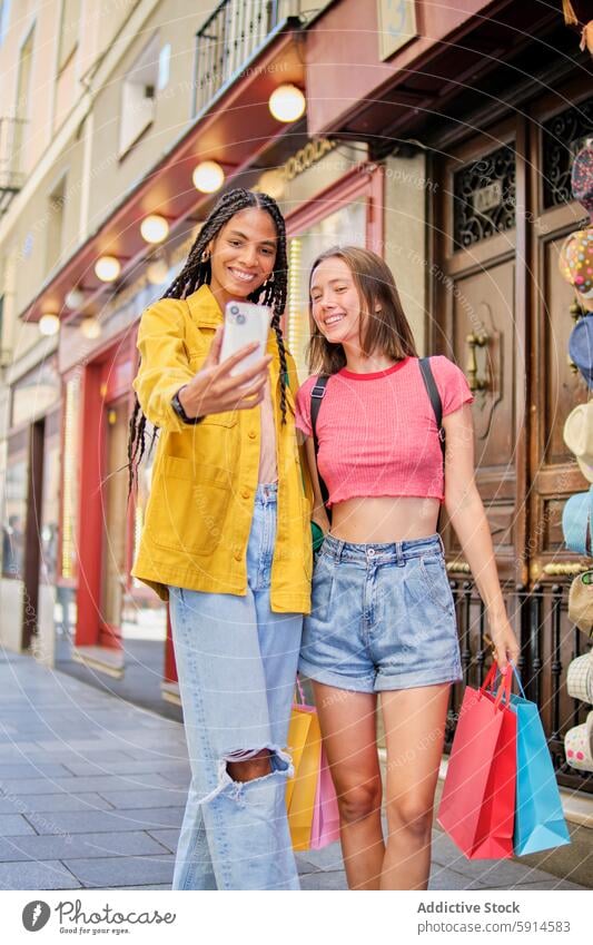 Two young multiethnic women enjoying shopping and taking a selfie female smartphone bag cheerful smile city street fashion youth friendship together technology