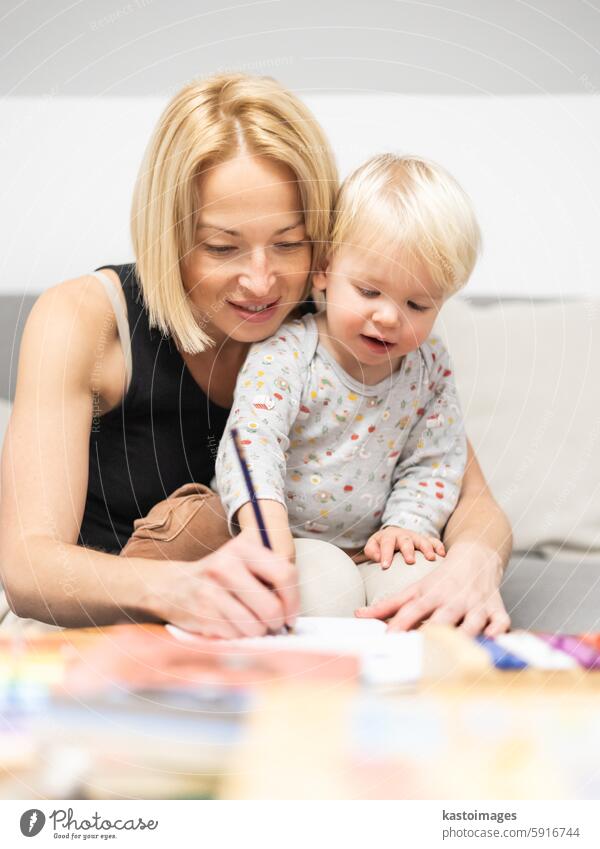 Caring young Caucasian mother and small son drawing painting in notebook at home together. Loving mom or nanny having fun learning and playing with her little 1,5 year old infant baby boy child.