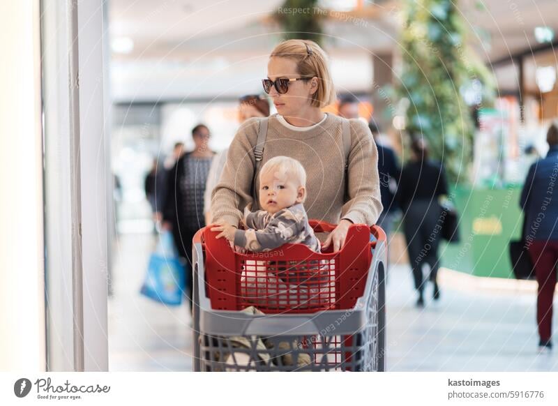 Mother shopping with his kid in the mall. Mother pushing shopping cart with her toddler baby boy child sitting in it along mall corridor. parent customer mom