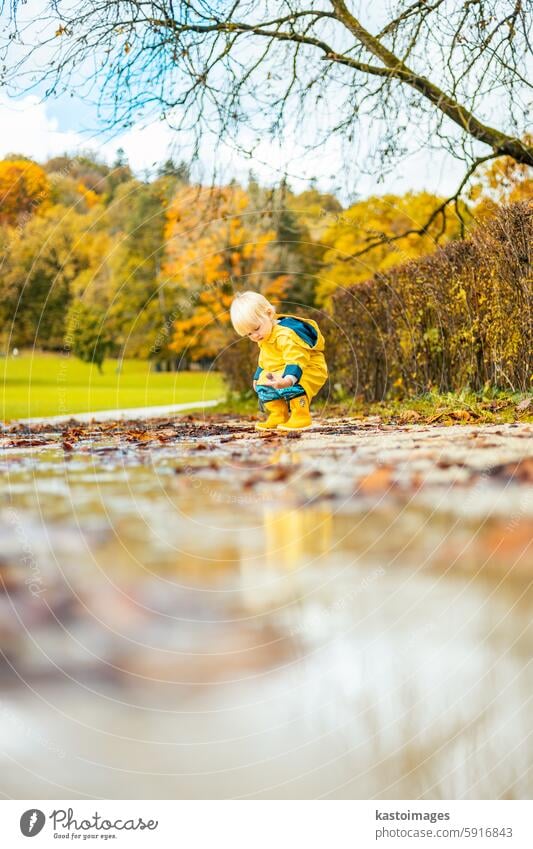 Sun always shines after the rain. Small bond infant boy wearing yellow rubber boots and yellow waterproof raincoat walking in puddles in city park on sunny rainy day.