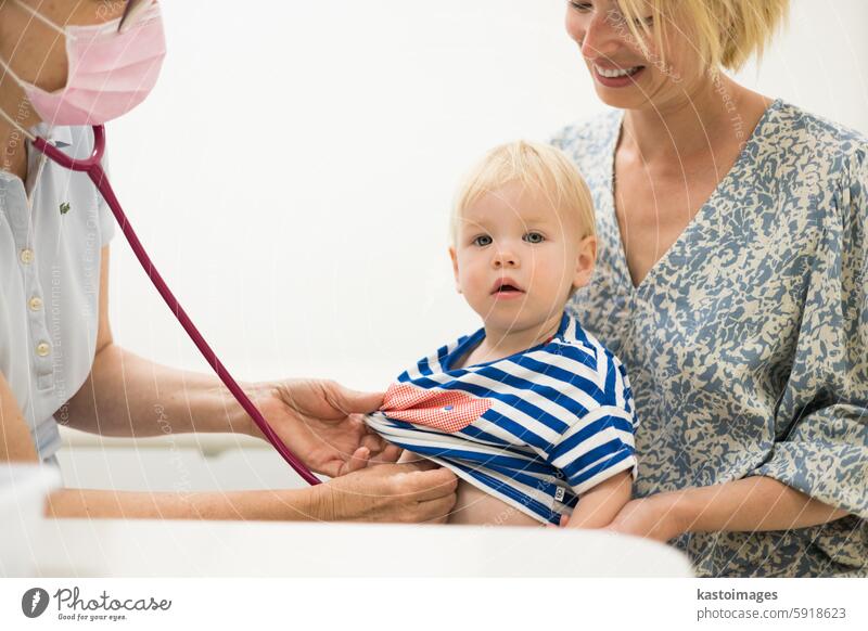 Infant baby boy child being examined by his pediatrician doctor during a standard medical checkup in presence and comfort of his mother. National public health and childs care care koncept.