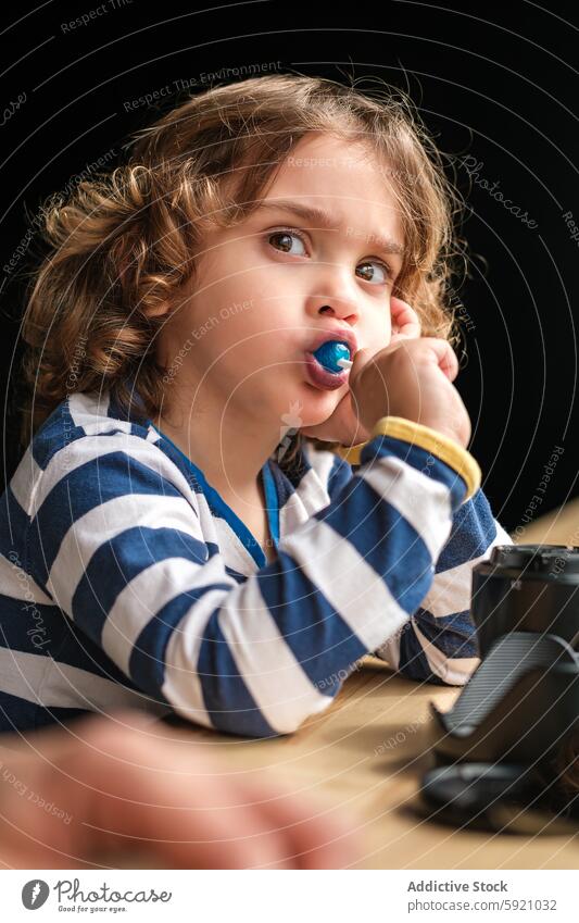 Young child savoring a blue lollipop enthusiastically curiosity girl camera lens candy mouth striped shirt play photography equipment black background toddler