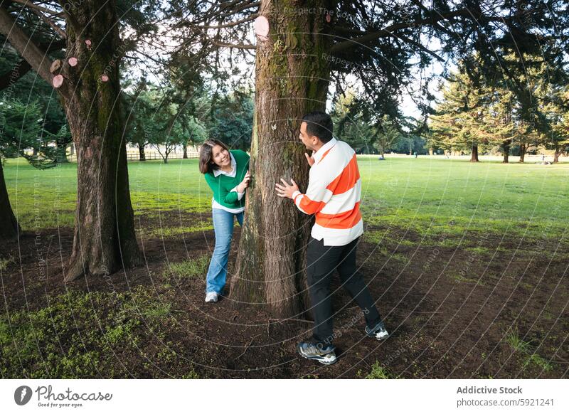 Young Latin couple enjoying a playful moment in park latin couple young woman peeking tree searching lush grass outdoors leisure casual clothing recreation