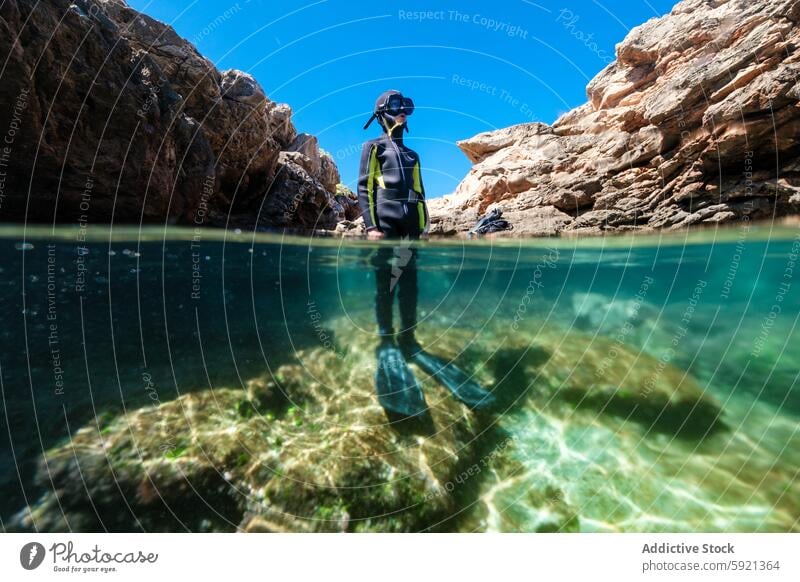 Unrecognizable teenage boy in a wetsuit and snorkel standing in the clear waters with rocky cliffs of Menorca, Spain, split view above and below the surface