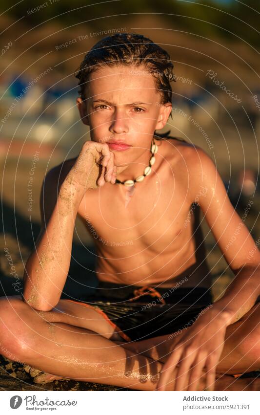 Thoughtful wet shirtless preteen boy wearing shell necklace sitting on sandy beach and looking at camera during summer vacation looking away thoughtful serious