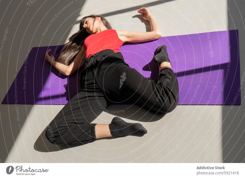 Woman relaxing in artistic pose on purple yoga mat woman relaxation sunlight shadow dynamic indoor red top black pant dance movement serene calm floor exercise