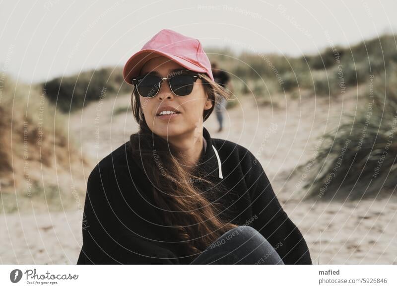 Young woman with sunglasses and baseball cap sitting on the beach Woman youthful Beach dunes Sunglasses View into the distance portrait Adults Exterior shot