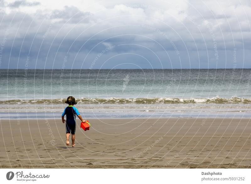 Small Boy Running Towards The Ocean on a Tropical Beach boy small small boy child small child hat sun hat swimming costume bucket pail run running beach sand