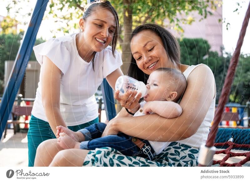 Baby being bottle-fed by two lesbian colombian women outdoors in a park setting baby bottlefeeding couple nurture care child infant tree playground swing