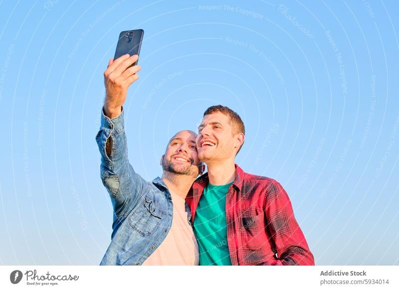 Gay couple enjoying time together while taking a selfie gay couple smartphone love relationship happiness men outdoor blue sky day smiling casual clothing