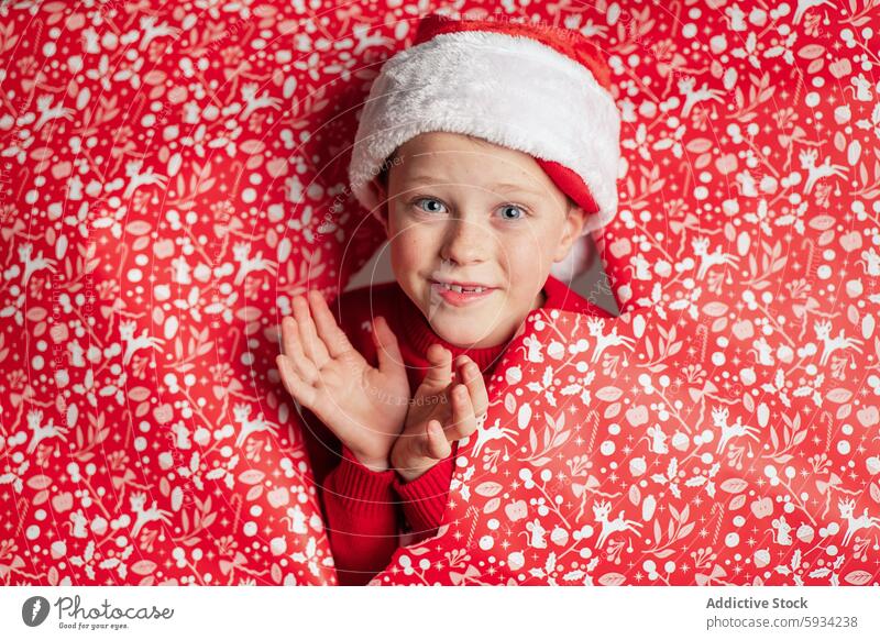 Young boy in Santa hat smiling amid red Christmas patterns christmas santa hat cheerful festive holiday playful background decoration seasonal joy clap hands