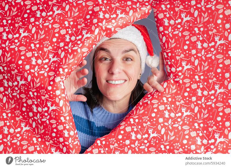 Woman peeking through Christmas wrapping paper, festive mood woman christmas santa hat holiday cheerful animal print red winter celebration joy fun pattern