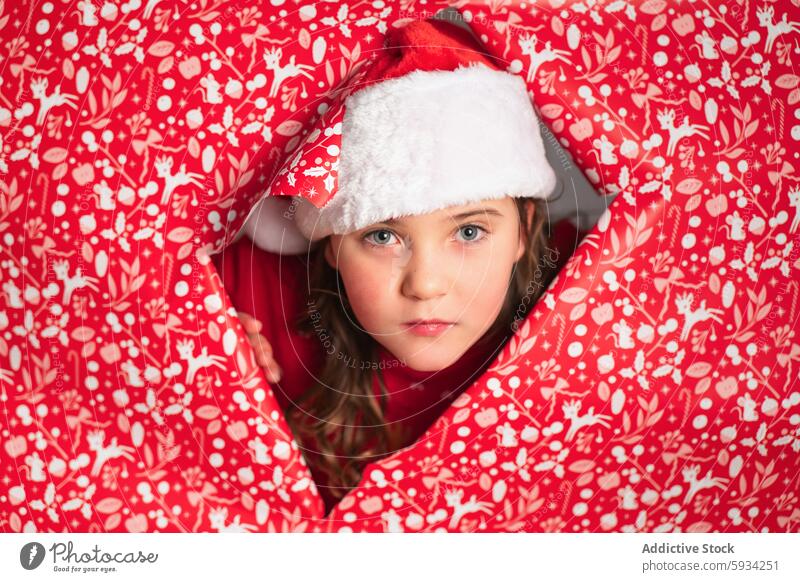 Young girl in Santa hat peeks out from red festive wrap christmas santa hat pattern holiday child serene expression youth cheerful celebration winter december